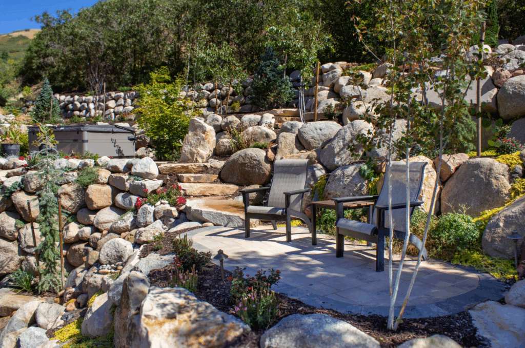 A terraced Utah backyard features large boulders used for retaining walls, a natural stone staircase, and a cozy seating area surrounded by trees, flowers, and landscape plants.